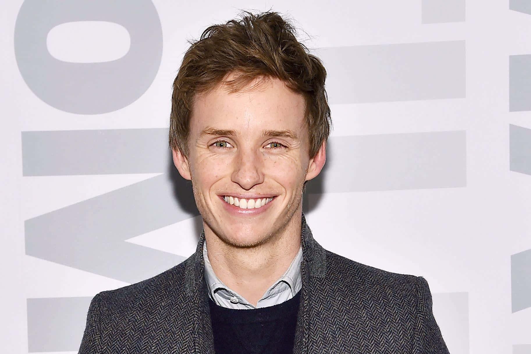 Eddie Redmayne Smiling In A Black Suit Against A Maroon Background Wallpaper