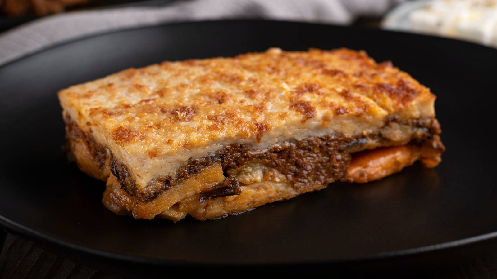 Slice Of Baked Moussaka On A Black Plate Wallpaper