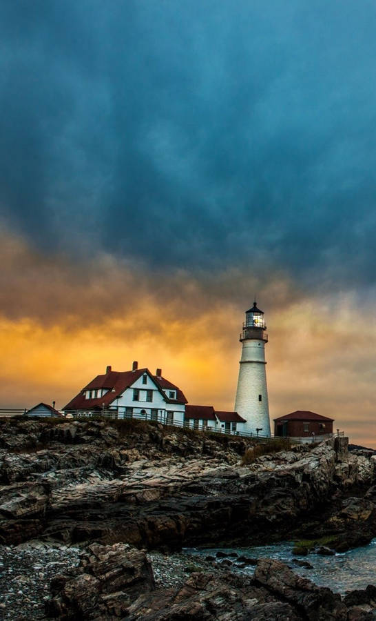 A Lighthouse Sits On A Rocky Shore With A Cloudy Sky Wallpaper