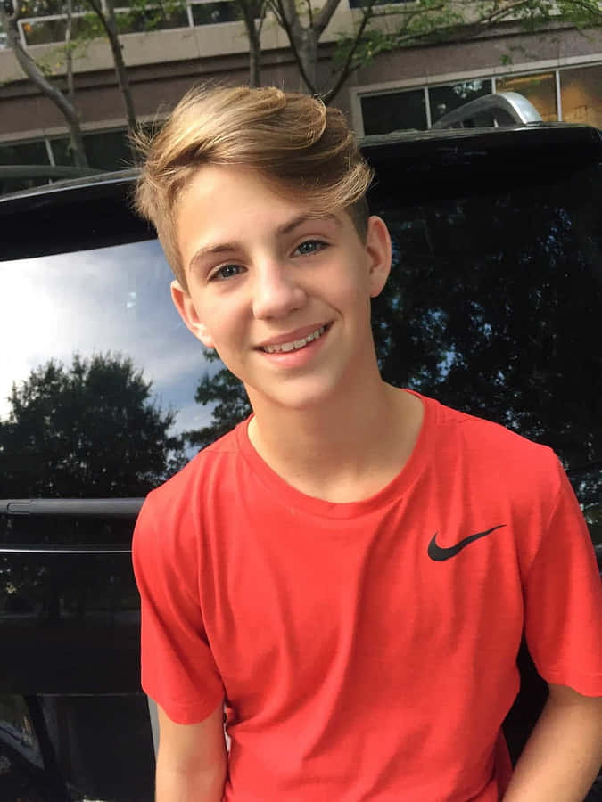 A Young Boy In A Red Shirt Smiling In Front Of A Car Wallpaper