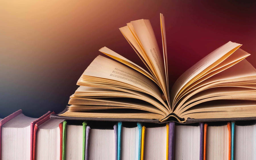 Opened Book Laying On Top Of A Stack Of Multicolored Books Wallpaper