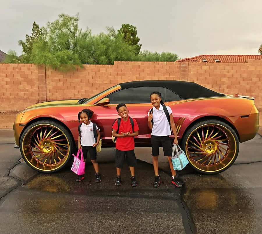 Three Children Standing Next To A Car With Large Wheels Wallpaper