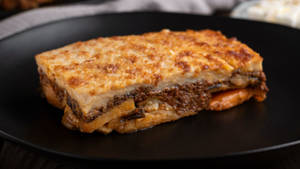 Slice Of Baked Moussaka On A Black Plate Wallpaper